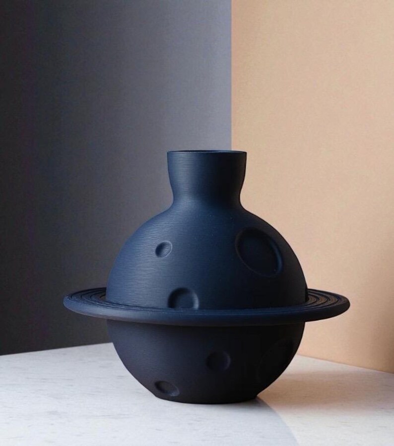 Może przedstawiać: Granatowy ceramiczny wazon o unikalnym planetarnym designie. Wazon ma kulistą podstawę z pierścieniem i cylindryczną g&oacute;rą. Powierzchnia ma kilka okrągłych wgłębień, przypominających kratery.
