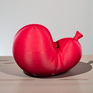 May include: A red balloon, partially deflated, rests on a light-colored wooden surface. The balloon's shape is distorted, with a knot at the end. The background is a plain, light gray.