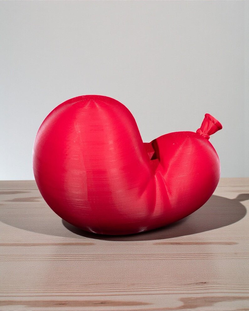 May include: A red balloon, partially deflated, rests on a light-colored wooden surface. The balloon's shape is distorted, with a knot at the end. The background is a plain, light gray.