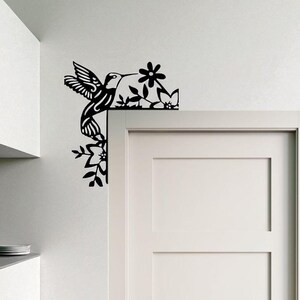 May include: Black metal hummingbird and floral corner decoration. The hummingbird is depicted with outstretched wings, feeding from a flower. The design is suitable for indoor or outdoor use and adds a decorative touch to any space.