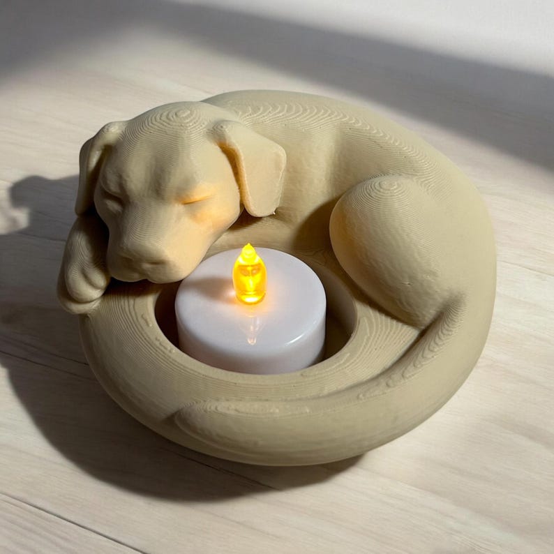 Puede incluir: Un portavelas en forma de perro de color beige con un dise&ntilde;o de perro durmiendo. El perro est&aacute; acurrucado alrededor de una vela blanca con una llama LED amarilla parpadeante. El portavelas est&aacute; hecho de un material texturizado.