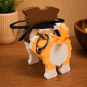 May include: A dog-shaped eyeglasses holder in shades of brown, orange, white, and black. The holder has a slot to hold eyeglasses and a ring to hold keys. The eyeglasses are black-framed. The holder is on a wooden surface.