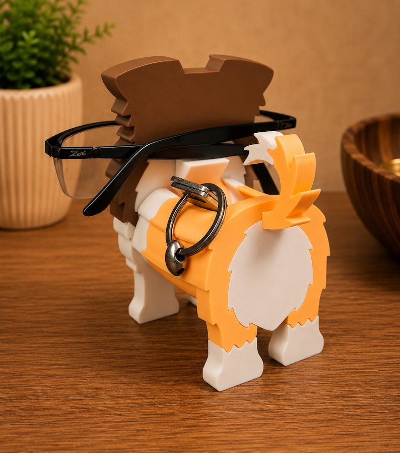 May include: A dog-shaped eyeglasses holder in shades of brown, orange, white, and black. The holder has a slot to hold eyeglasses and a ring to hold keys. The eyeglasses are black-framed. The holder is on a wooden surface.