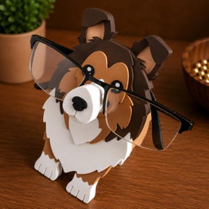 May include: A dog-shaped eyeglass holder in shades of brown, white, and tan, with black-framed glasses resting on its nose. The holder is made of layered materials, creating a 3D effect. The dog's features include a white muzzle and paws.