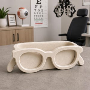 Op de afbeelding: Een beige brilvormige bureau-organizer. De organizer heeft een rechthoekige basis met een bril als hoofdontwerpelement. De achtergrond omvat een oogkaart, een oogmodel en optometrie-apparatuur.