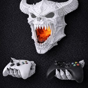 May include: A white, horned skull with a fiery interior, flanked by a white and a black video game controller, each held by a skeletal hand. The controllers have colored buttons. The items are displayed against a dark, textured background.
