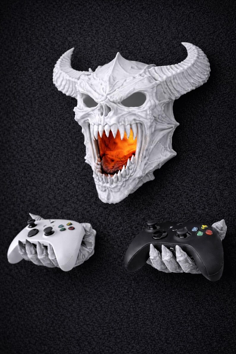 May include: A white, horned skull with a fiery interior, flanked by a white and a black video game controller, each held by a skeletal hand. The controllers have colored buttons. The items are displayed against a dark, textured background.