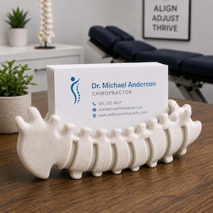 Op de afbeelding: Een cr&egrave;mekleurige, ruggengraatvormige visitekaarthouder met een stapel witte kaarten. De kaarten tonen de naam "Dr. Michael Anderson, Chiropractor" en contactgegevens. De houder staat op een houten oppervlak.