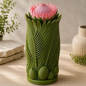 May include: A green vase with a detailed leaf design, holding a pink protea flower. The vase has a textured surface and a wide base. The flower is a vibrant pink with a spiky appearance, contrasting with the green of the vase.