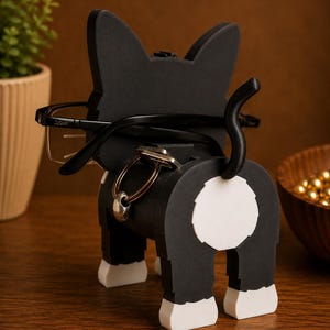 May include: A black and white cat-shaped eyeglasses and key holder. The holder has a black body with white accents on the paws and rear. Eyeglasses are placed on the cat's ears, and keys hang from the body.