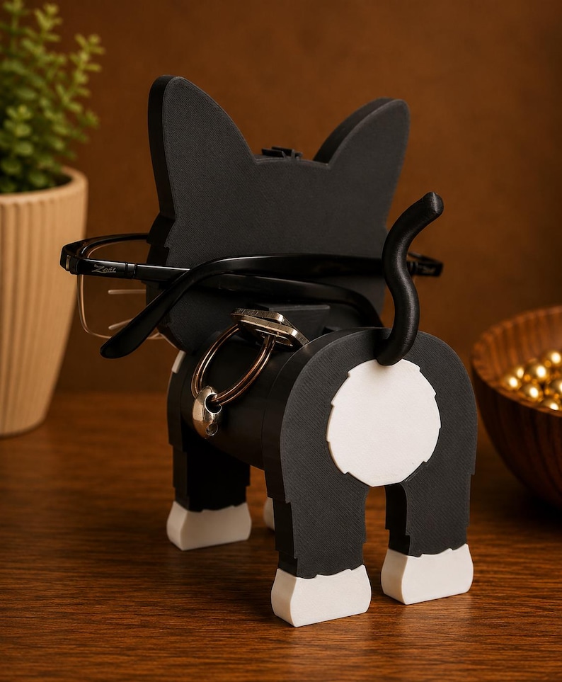 May include: A black and white cat-shaped eyeglasses and key holder. The holder has a black body with white accents on the paws and rear. Eyeglasses are placed on the cat's ears, and keys hang from the body.