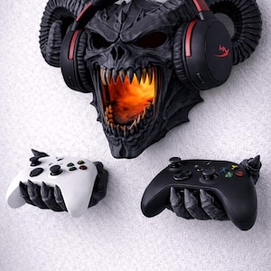 Puede incluir: Un conjunto de accesorios de juego montados en la pared en negro y gris. Presenta una calavera demon&iacute;aca negra con una boca naranja brillante, unos auriculares negros y dos soportes para mandos, uno blanco y otro negro.