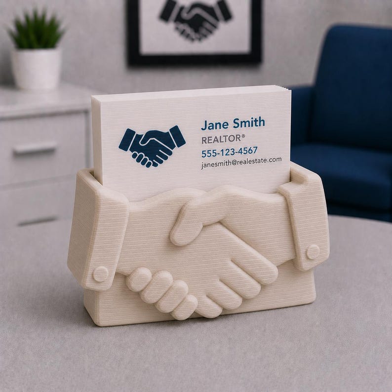 May include: A beige business card holder shaped like clasped hands, holding a stack of white business cards. The cards display a blue handshake logo, the name "Jane Smith, REALTOR&reg;", a phone number, and an email address.