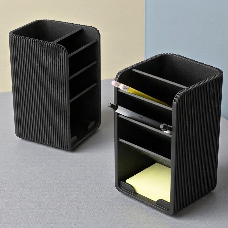 May include: Two black desk organizers with multiple compartments. One organizer has a pencil and pen in it, and the other has a yellow sticky note pad. The organizers have a ribbed design.