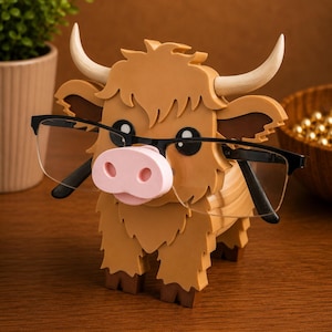 May include: A brown and tan Highland cow-shaped eyeglasses holder with a pink nose and black framed glasses. The holder has a shaggy fur design and curved horns. The glasses are held in place by the cow's nose.