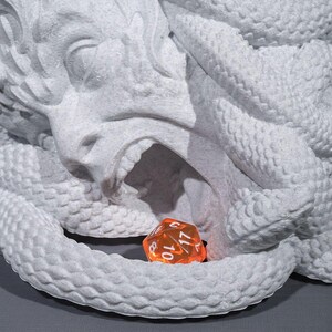 Peut inclure: Une sculpture d&eacute;taill&eacute;e de la t&ecirc;te d'une cr&eacute;ature mythique avec une bouche ouverte et des traits de serpent. Un d&eacute; orange &agrave; plusieurs faces repose &agrave; l'int&eacute;rieur de la bouche. La sculpture est de couleur gris clair.