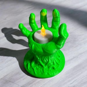 May include: A green, hand-shaped candle holder with a round base. The fingers of the hand cradle a white candle with a flickering yellow light. The candle holder is made of a textured material.