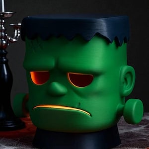 May include: A green Frankenstein's monster-shaped lamp with glowing orange eyes and mouth. The lamp has a black base and a dark blue top, with a jagged edge mimicking the monster's hair. A Halloween-themed decorative item.