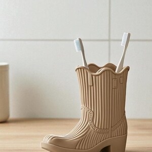 May include: A tan-colored toothbrush holder shaped like a cowboy boot, holding two white toothbrushes. The boot has a ridged texture and decorative stitching details. The holder sits on a wooden surface, with a neutral-toned background.