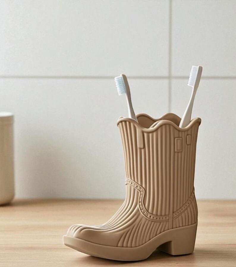 May include: A tan-colored toothbrush holder shaped like a cowboy boot, holding two white toothbrushes. The boot has a ridged texture and decorative stitching details. The holder sits on a wooden surface, with a neutral-toned background.