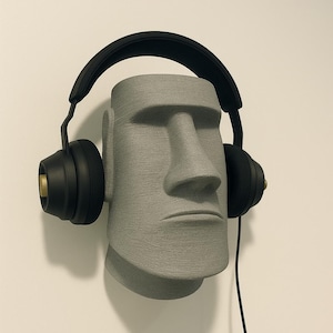 May include: A gray, stone-like sculpture of a Moai head wearing black headphones. The headphones have gold accents on the earcups and a black cable. The sculpture is mounted on a white wall.