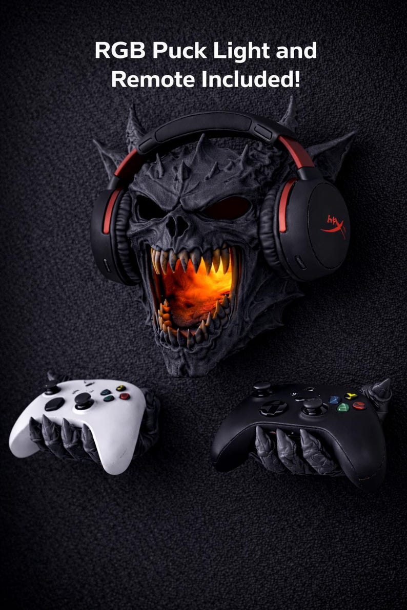May include: A dark, textured wall decor featuring a demonic skull with glowing orange light emanating from its mouth. A black headset with red accents rests on the skull. Two game controllers, one white and one black, are positioned below. Text reads: "RGB Puck Light and Remote Included!"