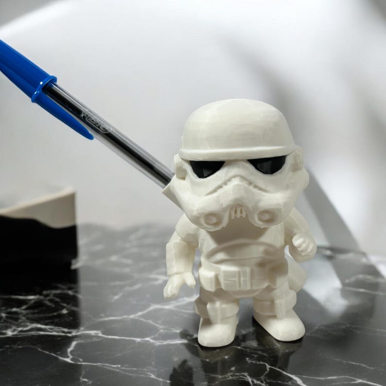 May include: A white Stormtrooper figurine with black eye lenses, standing upright. The figurine is detailed with a helmet, armor, and a blaster. A blue and silver pen is positioned near the figurine.