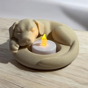 Puede incluir: Un portavelas beige con forma de perro con un dise&ntilde;o de perro durmiendo. El perro est&aacute; acurrucado alrededor de una vela blanca con una llama LED amarilla. El portavelas es de madera.