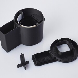 Op de afbeelding: Zwarte plastic auto-organizer met een bekerhouder, een klein vak en een verwijderbare telefoonhouder. De telefoonhouder heeft het label "Phone Dock".