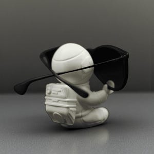 May include: A white astronaut figurine with a backpack, seated and wearing black sunglasses. The figurine is set against a gray background, creating a minimalist aesthetic. The sunglasses are positioned as if the astronaut is wearing them.