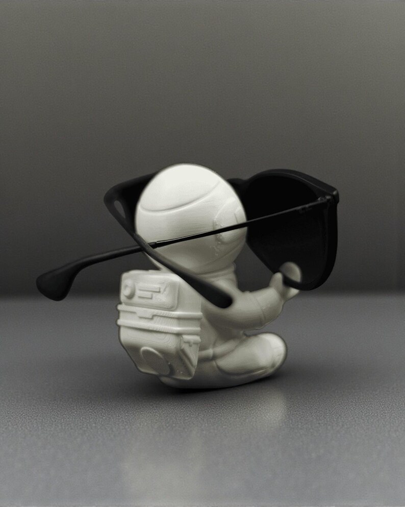 May include: A white astronaut figurine with a backpack, seated and wearing black sunglasses. The figurine is set against a gray background, creating a minimalist aesthetic. The sunglasses are positioned as if the astronaut is wearing them.