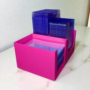 May include: A bright pink desk organizer with multiple compartments. The organizer holds stacks of blue cards. The design features a stepped structure with a rectangular base and a curved side for easy access to the cards.