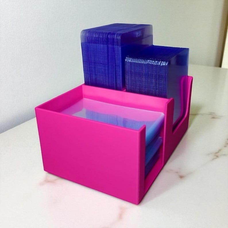 May include: A bright pink desk organizer with multiple compartments. The organizer holds stacks of blue cards. The design features a stepped structure with a rectangular base and a curved side for easy access to the cards.