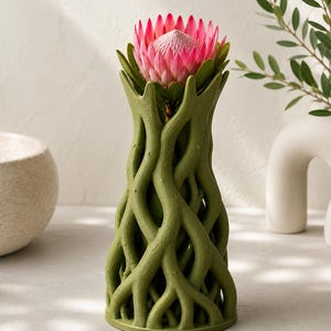 May include: A green, tree-trunk-shaped vase holds a vibrant pink protea flower. The vase has a textured, organic design with interwoven branches. The flower's petals are a deep pink, contrasting with the green of the vase and leaves.