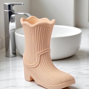 May include: A peach-colored decorative boot-shaped vase with a ridged texture. The vase has a scalloped top, a small heel, and a strap detail. The boot is set against a bathroom background with a white sink and chrome faucet.