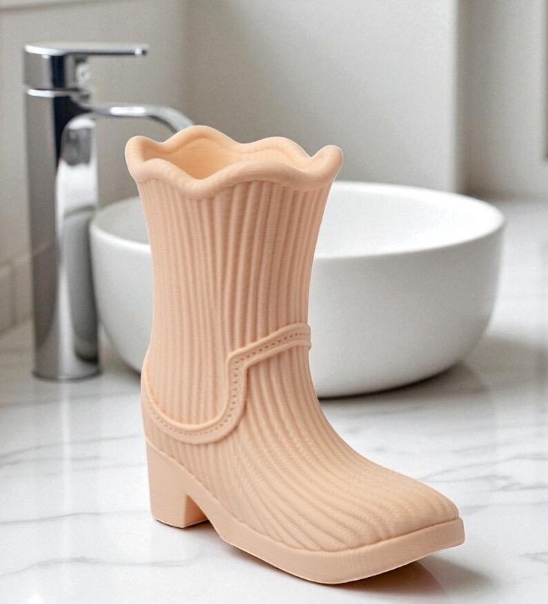 May include: A peach-colored decorative boot-shaped vase with a ridged texture. The vase has a scalloped top, a small heel, and a strap detail. The boot is set against a bathroom background with a white sink and chrome faucet.