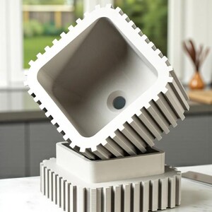 May include: A modern, gray, square sink with a unique, ridged design. The sink is elevated on a matching base, creating a striking visual. The interior of the sink features a drain hole. The overall aesthetic is minimalist and contemporary.