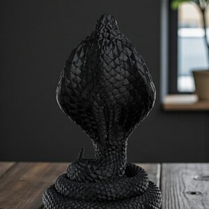 Op de afbeelding: Zwarte cobra-sculptuur met een textuur, schubachtig patroon. De cobra is opgerold en rechtop, met zijn kap uitgespreid. De sculptuur is gemaakt van een donker materiaal en staat op een houten oppervlak.