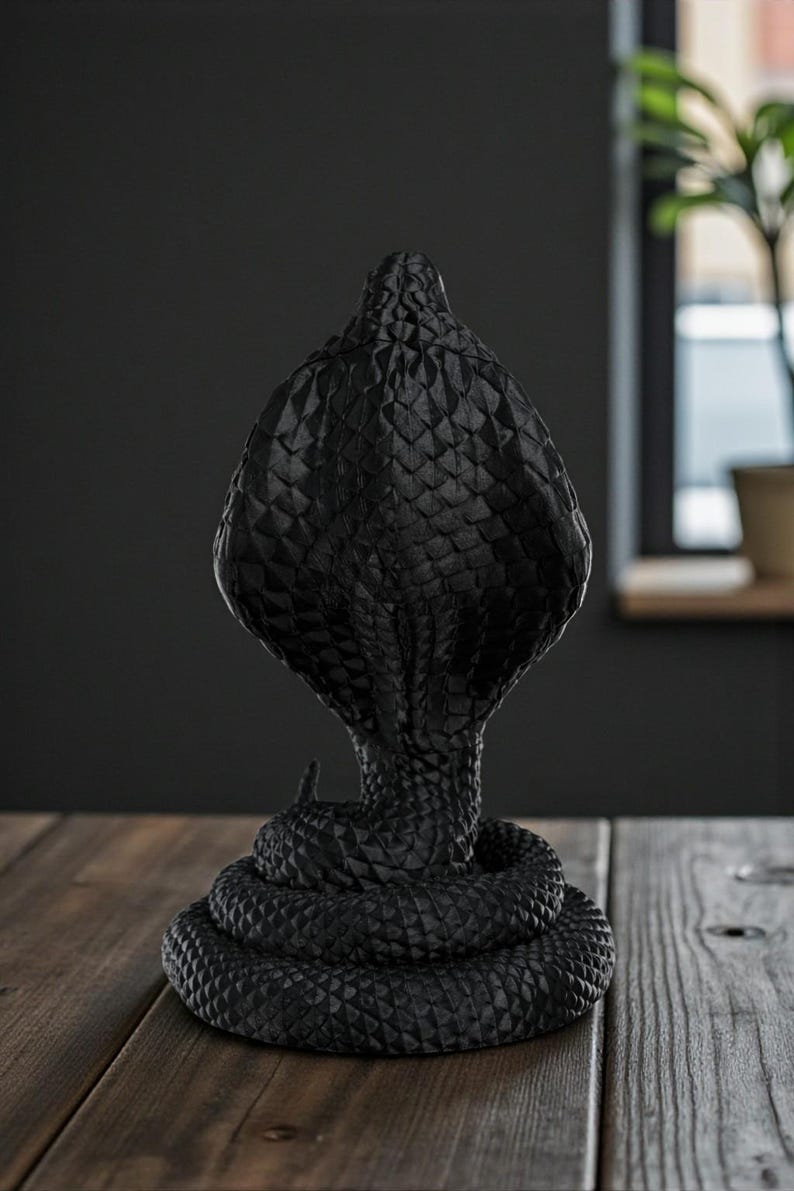 Op de afbeelding: Zwarte cobra-sculptuur met een textuur, schubachtig patroon. De cobra is opgerold en rechtop, met zijn kap uitgespreid. De sculptuur is gemaakt van een donker materiaal en staat op een houten oppervlak.