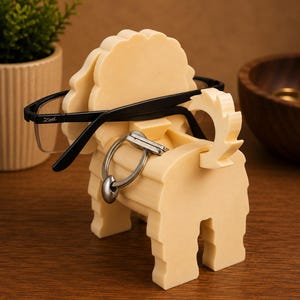 May include: A cream-colored, dog-shaped eyeglasses holder with black-framed glasses resting on it. A silver key ring hangs from the holder. The holder is made of a light-colored material and sits on a wooden surface.