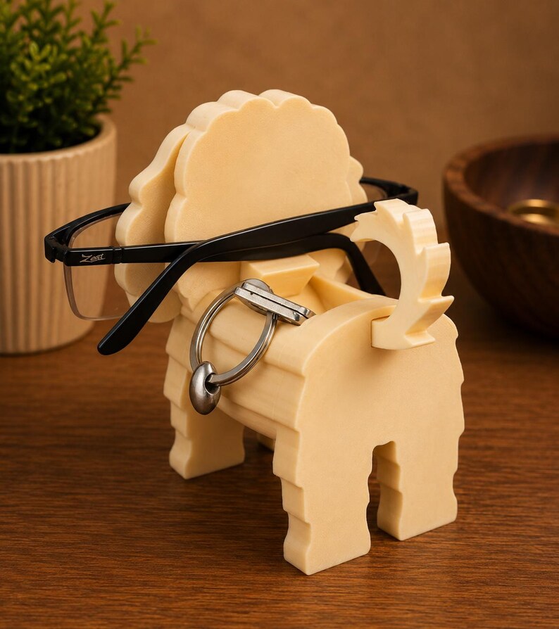 May include: A cream-colored, dog-shaped eyeglasses holder with black-framed glasses resting on it. A silver key ring hangs from the holder. The holder is made of a light-colored material and sits on a wooden surface.