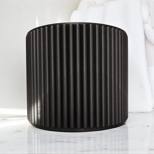 May include: A black, cylindrical container with vertical, ridged detailing. The container is made of a matte material and sits on a white marble surface. White towels are in the background.