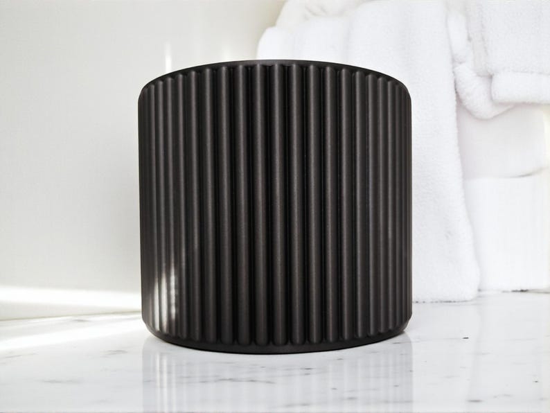 May include: A black, cylindrical container with vertical, ridged detailing. The container is made of a matte material and sits on a white marble surface. White towels are in the background.
