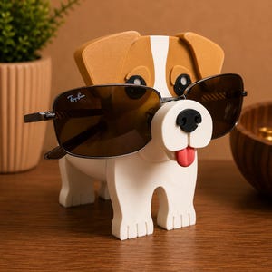 May include: A whimsical dog-shaped eyeglass holder in white, brown, and black. The holder has a red tongue and a black nose. A pair of Ray-Ban sunglasses with dark lenses rests on the holder. The holder is on a wooden surface.