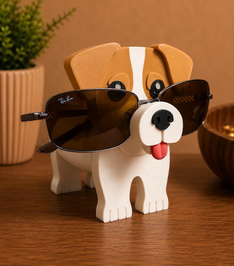 May include: A whimsical dog-shaped eyeglass holder in white, brown, and black. The holder has a red tongue and a black nose. A pair of Ray-Ban sunglasses with dark lenses rests on the holder. The holder is on a wooden surface.