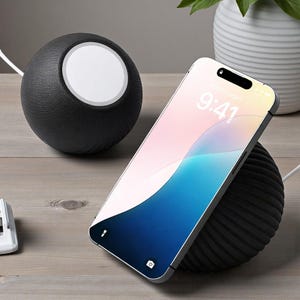 May include: A black wireless charging station with a matching phone stand. The phone displays the time as 9:41. The charging station has a textured, spherical design. A white cable connects the phone stand to the power source.