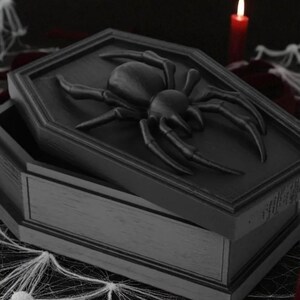 May include: A black coffin-shaped box with a large spider on the lid. The box is open, revealing the interior. The scene is set with a red candle and spider web decorations, creating a Halloween theme.