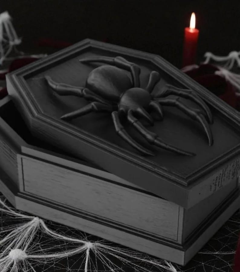 May include: A black coffin-shaped box with a large spider on the lid. The box is open, revealing the interior. The scene is set with a red candle and spider web decorations, creating a Halloween theme.