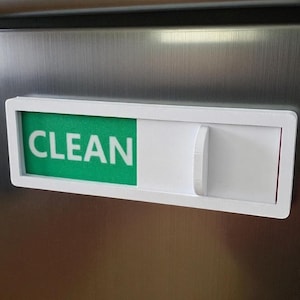 May include: A white plastic dishwasher indicator with the word "CLEAN" in white letters on a green background. The indicator has a sliding panel to display "CLEAN" or "DIRTY".