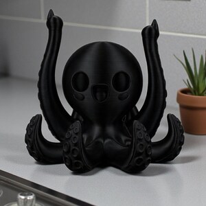 Puede incluir: Figura de pulpo negro, impresa en 3D, con una cara sonriente y tent&aacute;culos levantados. La figura est&aacute; sobre una superficie clara, con una peque&ntilde;a planta en maceta y el n&uacute;mero 25 en la pared. El pulpo tiene un dise&ntilde;o lindo y de dibujos animados.